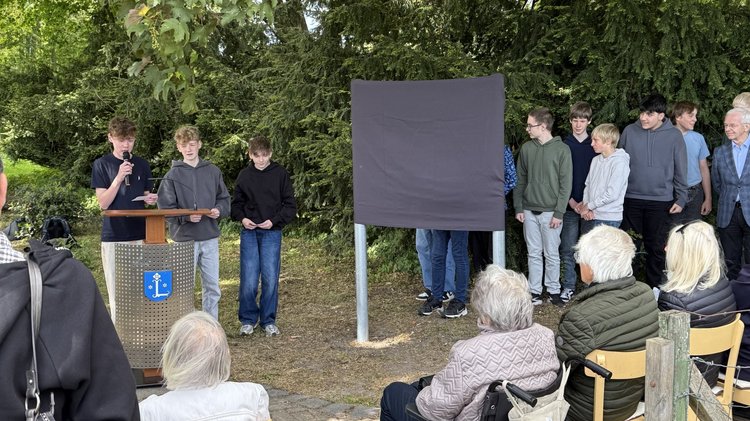 Links stehen drei Schüler am Rednerpult, einer spricht in ein Mikrofon. Rechts die verhüllte Tafel und weitere Jugendliche. Im Vordergrund einige zuhörende Gäste von hinten aufgenommen. 