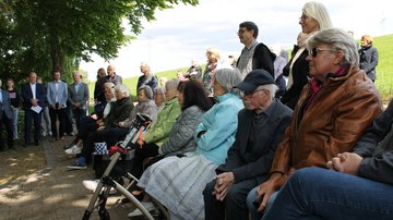 Mehrere Personen, teils sitzend, teils stehend, die nach links schauen. Sie hören einem Redner zu.